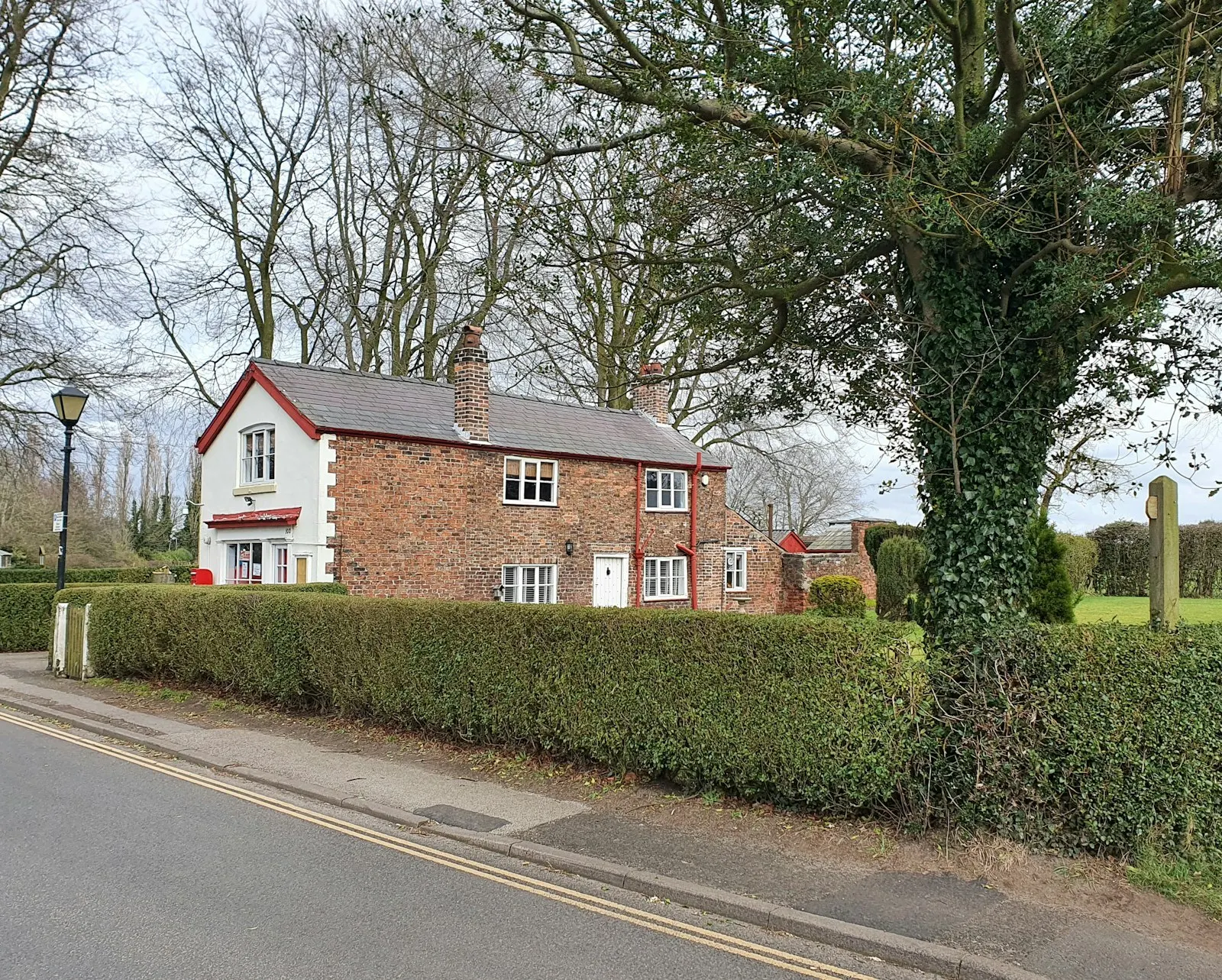 A small brick and white cottage behind a hedge on a village road