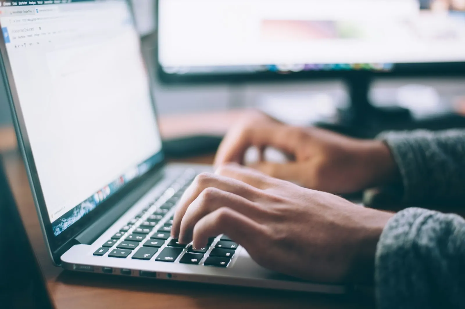 Hands typing on a laptop keyboard in front of a monitor