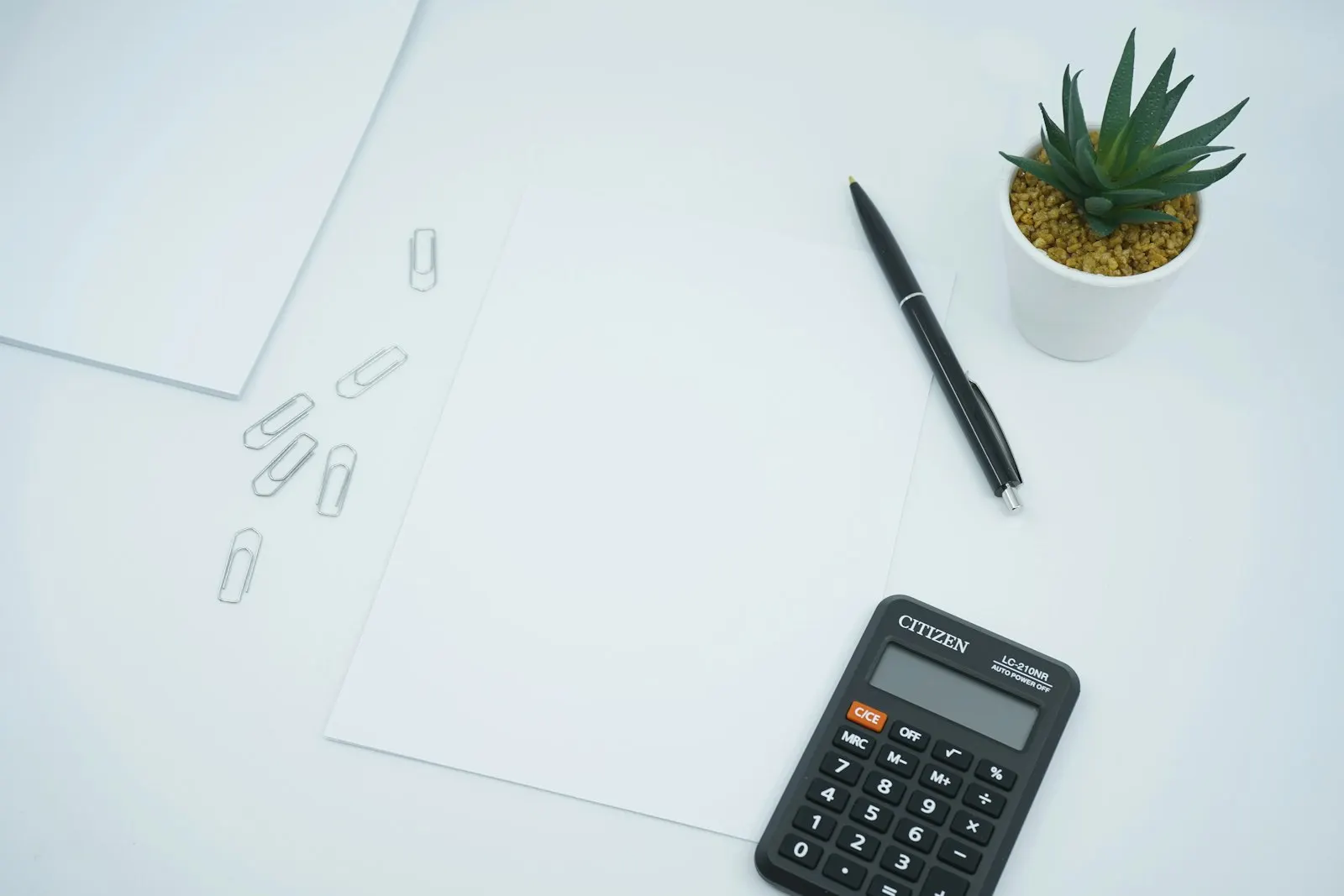 A calculator, blank sheets of paper, paperclips and a small plant on a pale desk