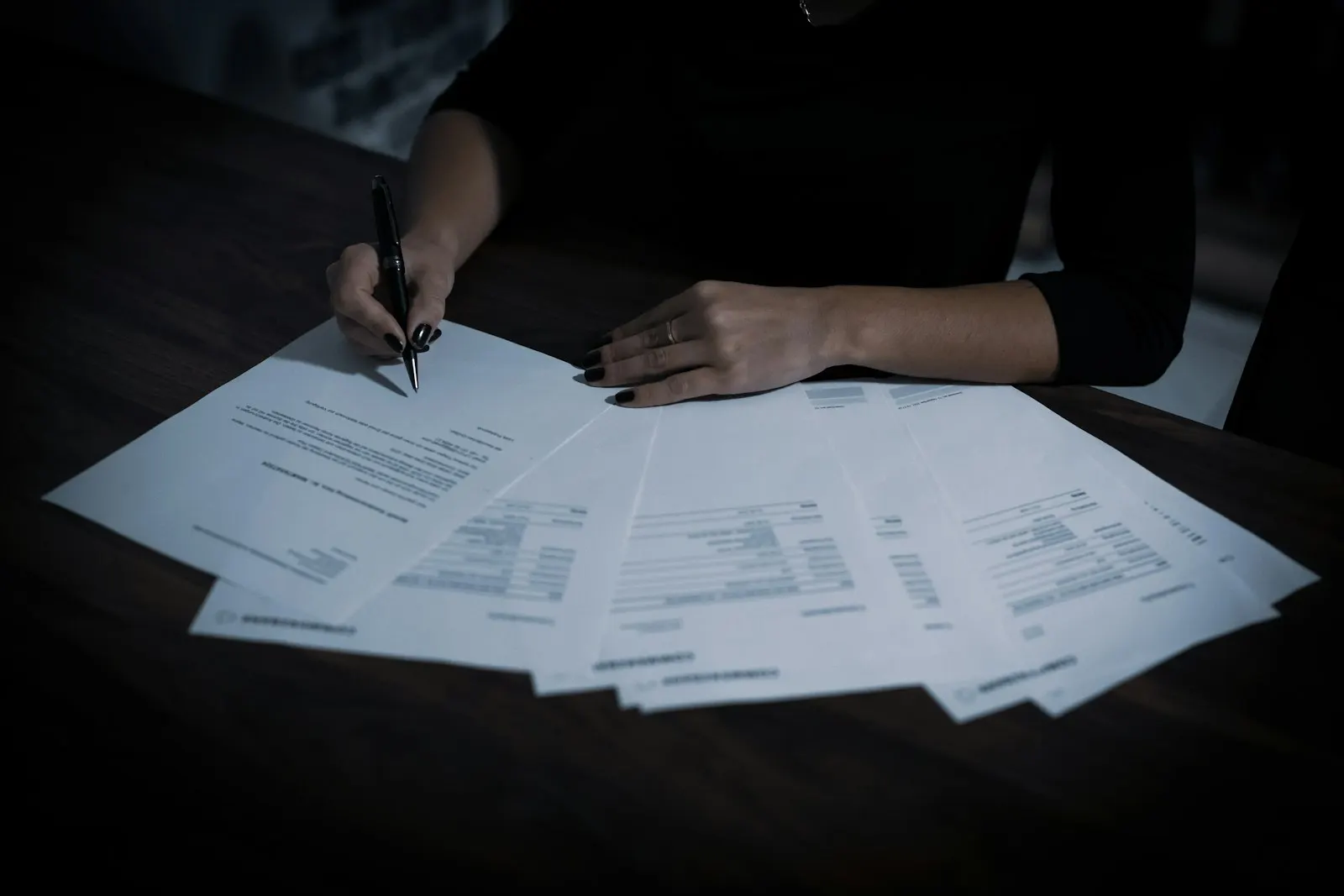 Hands filling in printed forms spread across a dark desk
